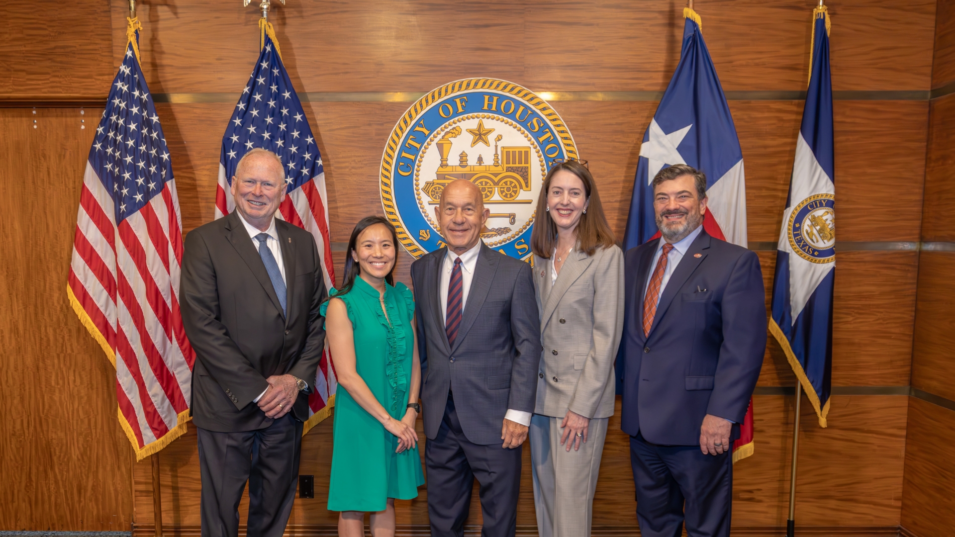 Five individuals stand side by side in front of a wood-paneled wall displaying the “City of Houston” seal. Two U.S. flags are positioned on the left, and the Texas state flag and a City of Houston flag are on the right.
